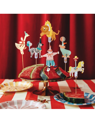 Topper de gâteau lion circus sur morceaux de pastèque décorative, entouré d’animaux en bois colorés sur pics, fond rouge.