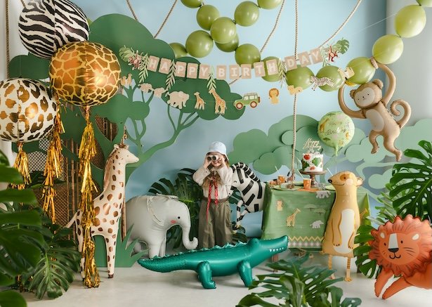 Décor de fête coloré avec ballons animaux et guirlande « Happy Birthday », enfant en costume safari entouré d’éléphants et d’un crocodile gonflable.