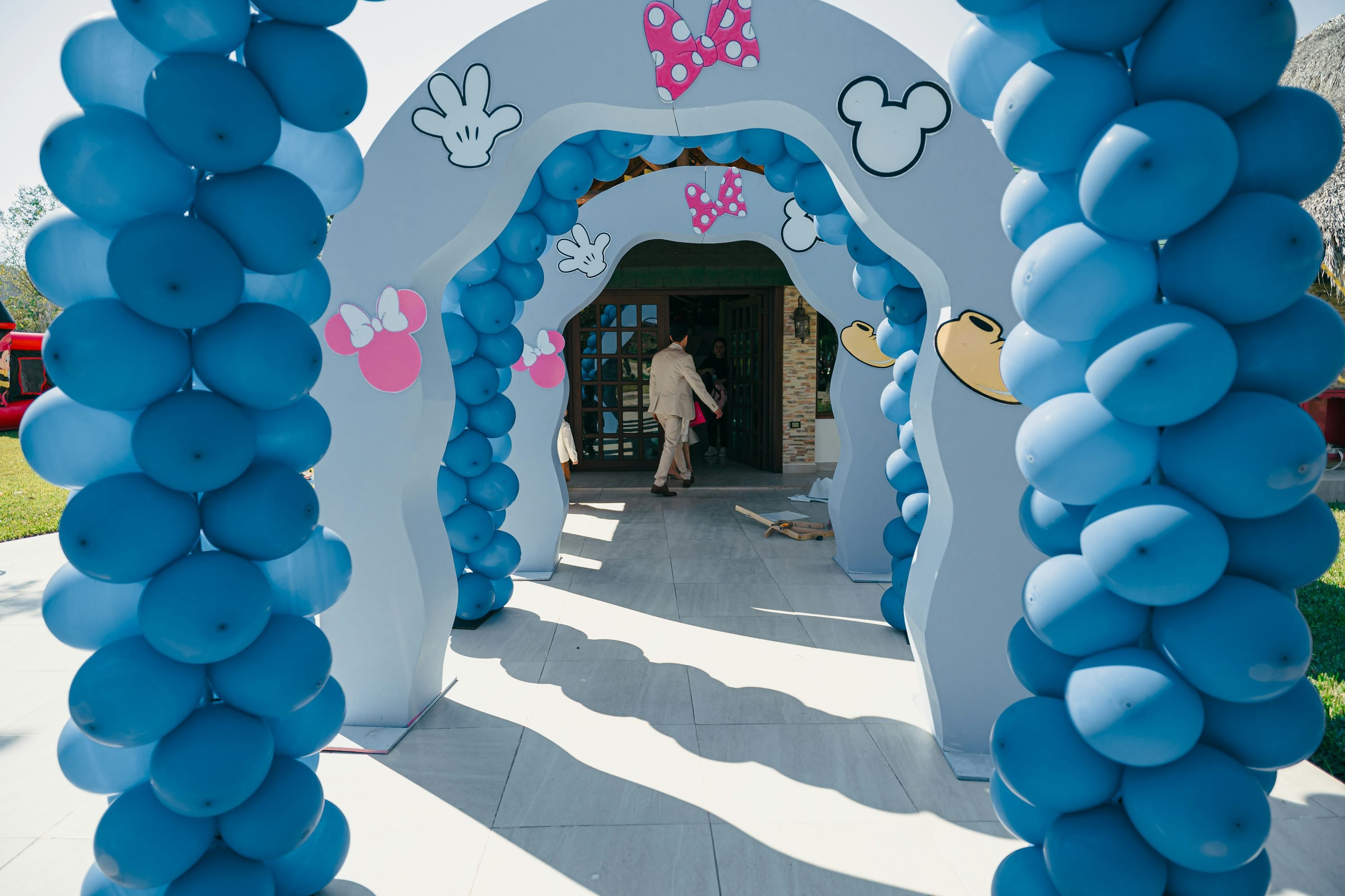 Entrée décorée avec des arches bleues en ballons formant un tunnel festif, motifs Minnie et Mickey, et silhouettes d’ombrelle; personnes entrant dans le bâtiment.