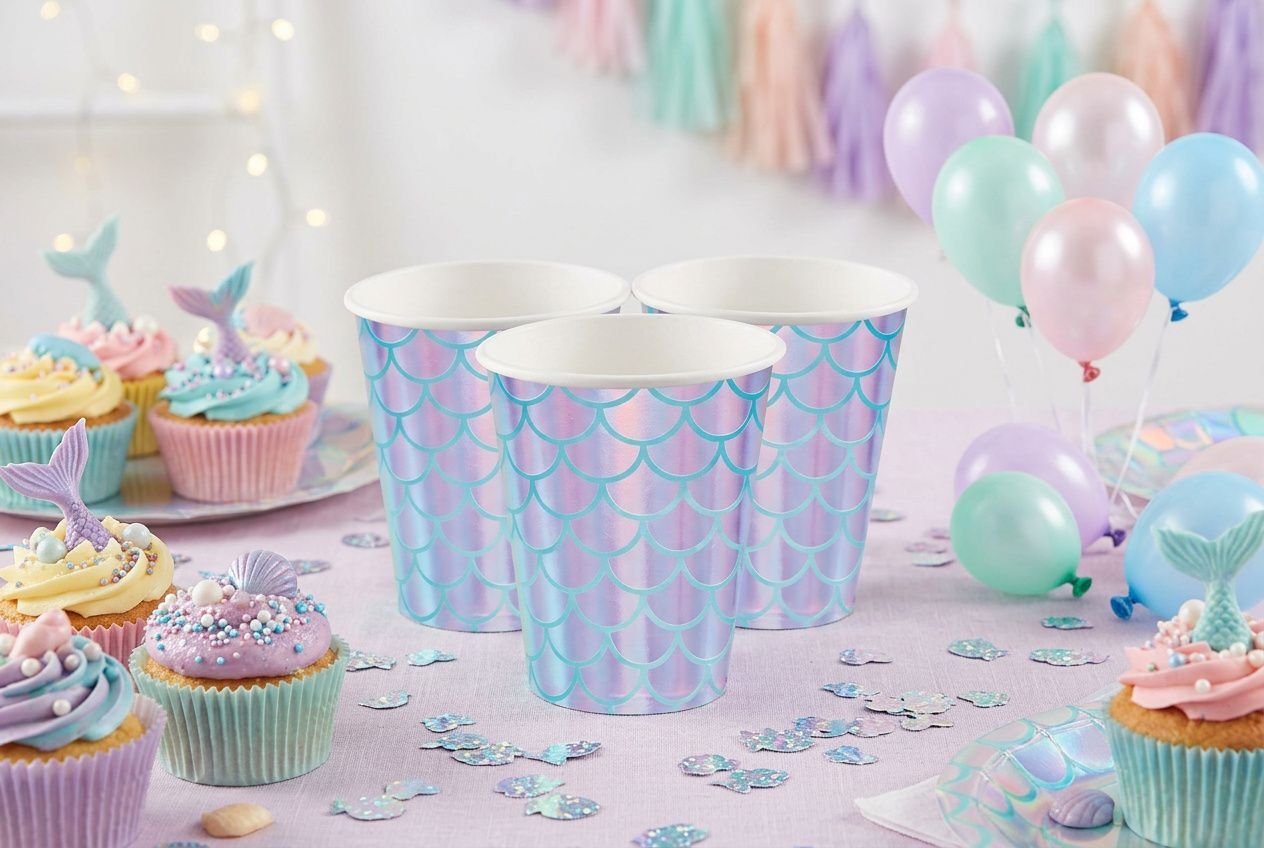 Gobelets jetables colorés fête en plastique bleu clair avec motif écaillé, entourés de cupcakes pastel et ballons.