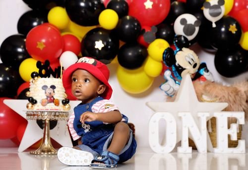 Petit garçon sur fond de décor de fête, devant un gâteau et des ballons colorés « ONE » en grandes lettres.